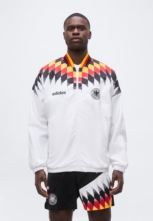 Άνδρας που φοράει λευκό μπουφάν Adidas και μαύρο σορτς με κόκκινο, κίτρινο και μαύρο μοτίβο ρόμβων και λογότυπα της Γερμανικής Ποδοσφαιρικής Ομοσπονδίας.