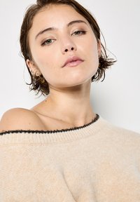 Suéter beige con escote asimétrico que presenta un cuello con ribete negro, textura de punto y un corte relajado, destacando una estética acogedora y casual.