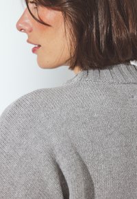 Jonge vrouw met kort bruin haar, gekleed in een lichtgrijze gebreide trui, die zijwaarts kijkt tegen een effen witte achtergrond.