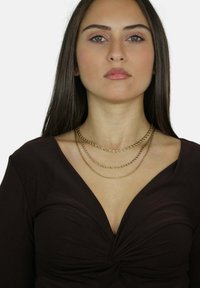 Collana in oro con tre strati sovrapposti, caratterizzata da maglie attorcigliate, indossata sopra una maglia scura e aderente con scollatura a V profonda e dettaglio arricciato.