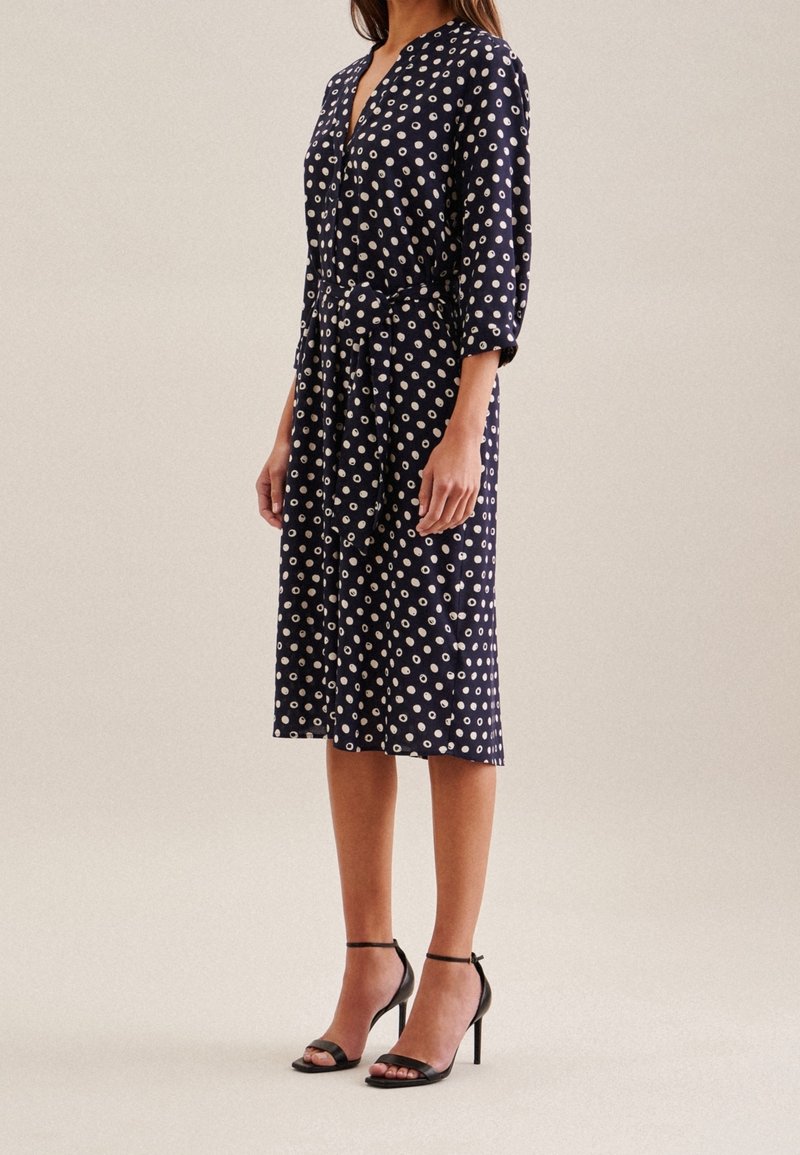 Femme portant une robe bleu marine longueur genou à pois blancs, manches trois-quarts, ceinture à la taille et sandales à talons hauts noires.