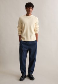 Jasnożółty sweter z prążkowanej dzianiny o luźnym kroju, w połączeniu z ciemnoniebieskimi, oversize'owymi spodniami i czarnymi loafersami. Prosta, codzienna stylizacja.