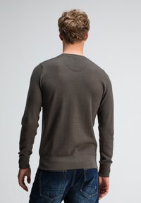 Mann mit kurzen, lockigen blonden Haaren, der von hinten steht, trägt einen strukturierten dunkelgrauen Pullover und blaue Jeans vor einem schlichten hellen Hintergrund.