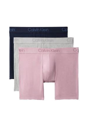 Uomo senza maglietta in piedi a piedi nudi su uno sfondo grigio uniforme, indossa boxer Calvin Klein rosa chiaro.