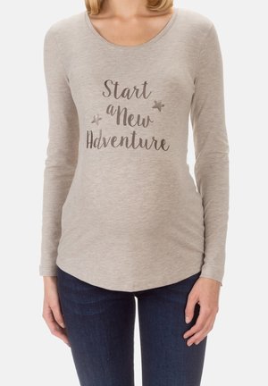 Kobieta nosząca beżową koszulę z długim rękawem z napisem "Start a New Adventure" i dwoma małymi grafikami gwiazdek, w połączeniu z ciemnoniebieskimi dżinsami.
