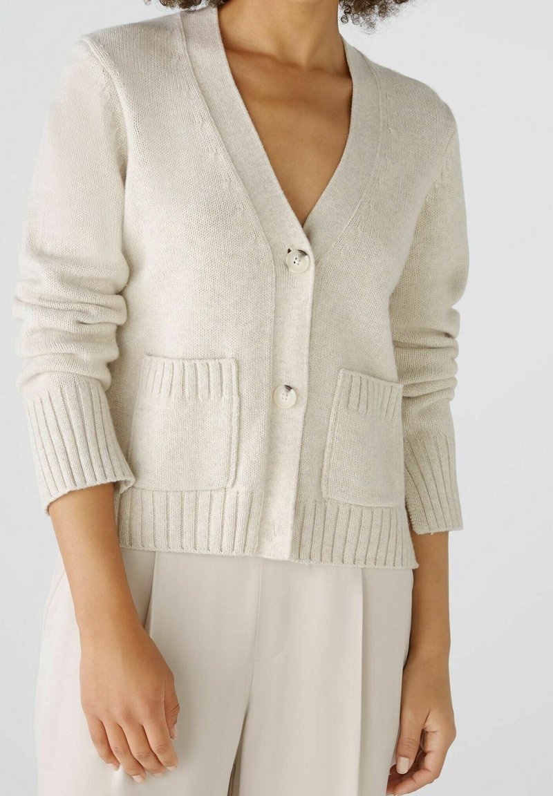 Femme portant un cardigan en maille crème avec deux poches avant et des boutons, associé à un pantalon beige clair, coupé au niveau du cou et des hanches.