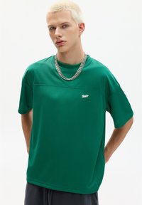 Tricou sportiv verde cu mâneci scurte, având o textură distinctivă și un panou cusut contrastant. Dispune de un mic logo alb pe piept.