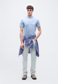 Νέος άνδρας που φοράει ανοιχτό μπλε μπλούζα Aston Martin, ανοιχτόχρωμο παντελόνι, γκρι μπότες και μπλε καρό πουκάμισο δεμένο γύρω από τη μέση, στέκεται με τα χέρια στις τσέπες.