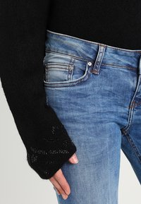 Suéter negro de punto con mangas anchas, combinado con jeans de mezclilla azul desgastados y ajustados que presentan costuras decorativas y diseño de cinco bolsillos.