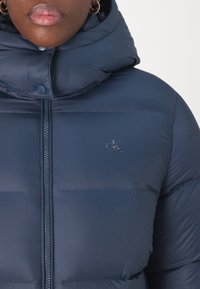 Gros plan sur une personne portant une veste rembourrée bleu marine avec un col haut, une fermeture éclair visible, un petit logo cK sur la poitrine et des boucles d'oreilles en forme de créole dorées.
