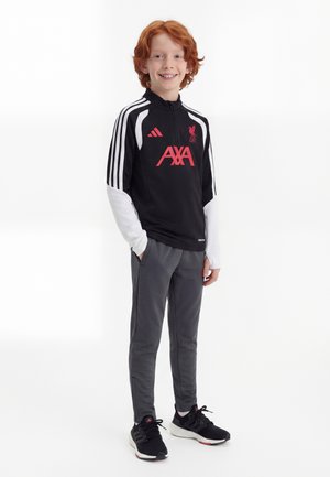 Uśmiechnięty rudowłosy chłopiec w czarno-białej sportowej kurtce Liverpool FC, szarych spodniach i czarnych butach Adidas, stojący z rękami w kieszeniach.
