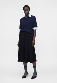 Femme portant un pull bleu marine par-dessus une chemise bleu clair à col, une jupe midi noire, des chaussettes grises et des chaussures à talons noires sur fond blanc.