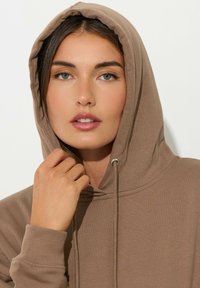 Felpa marrone con cappuccio e cordini, dal materiale morbido e con polsini a coste. Il modello ha i capelli lunghi e tiene la scollatura, guardando in avanti.