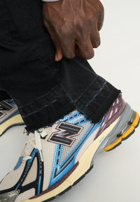 Zapatilla blanca y gris con acentos azules y detalles morados, combinada con vaqueros negros con bordes deshilachados en el dobladillo. Mano agarrando la pierna del pantalón.