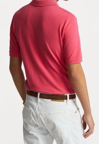 Kortärmad polo i klarröd, gjord av mjukt tyg. Bärs med vita byxor och ett brunt bälte, vilket visar en avslappnad stil.