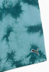 Tissu tie-dye bleu sarcelle et vert foncé avec un patch du logo Puma près de l'ourlet cousu.