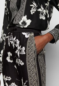 Tenue noire avec des motifs floraux et cachemire blancs, manches longues, pantalon à taille élastique, et une main placée dans la poche latérale.