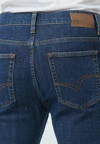 Donkerblauwe denim jeans met een rode en gele accentstiksel, voorzien van een leren patch en twee zichtbare achterzakken.