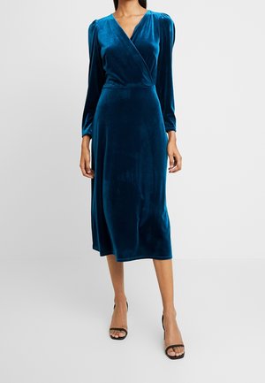Donna che indossa un abito avvolgente in velluto blu petrolio con maniche lunghe e lunghezza midi, abbinato a sandali neri con tacco alto e punta aperta.