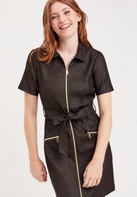 Robe noire à manches courtes avec fermeture éclair à l'avant et deux poches zippées dorées. Comprend une ceinture en tissu et présente un motif texturé subtil.