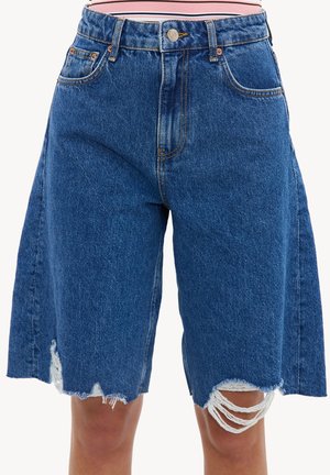 Short en jean bleu taille haute avec ourlet effiloché et détails déchirés porté par une personne debout devant un fond uni.