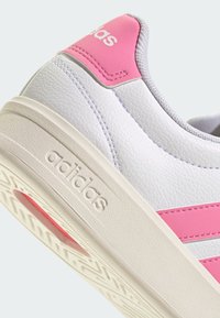 White sneaker with pink accents, textured leather upper, and beige sole. Features adidas logo, flat laces, and rubber tread pattern.