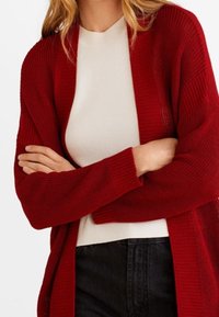 Femme portant un cardigan tricoté rouge profond sur un top blanc, les bras croisés, associé à un jean foncé taille haute.