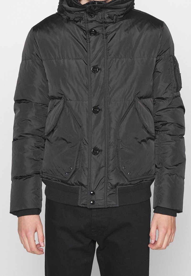 Black puffer jacket with a high collar, front snap buttons, and side pockets. Features a smooth, shiny texture and elastic cuffs.