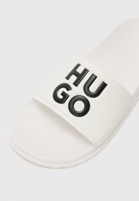 Witte schuif sandaal met een brede band met geperste zwarte letters die "HUGO" spellen. Gladde textuur met een gevormde voetbed en duurzame zool.