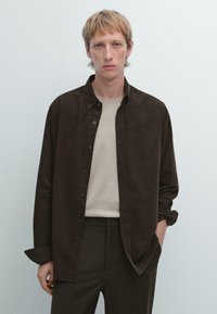 Braun gestreifte Hemdjacke aus weichem Stoff, getragen über einem hellbeigen Rundhals-T-Shirt, kombiniert mit dunklen Hosen. Das Modell hat kurze Haare und einen neutralen Hintergrund.