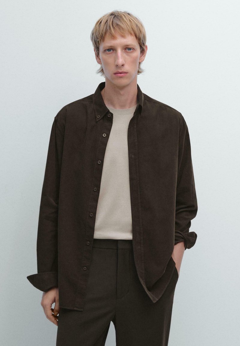 Braun gestreifte Hemdjacke aus weichem Stoff, getragen über einem hellbeigen Rundhals-T-Shirt, kombiniert mit dunklen Hosen. Das Modell hat kurze Haare und einen neutralen Hintergrund.