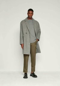 Šedý vlněný kabát s dvouřadým zapínáním, položený na šedé košili a olivově zelených kalhotách. Černé boty dotvářejí celý outfit.