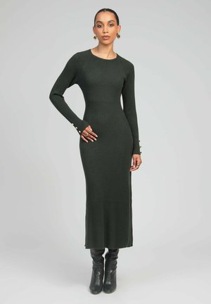 Robe en maille côtelée vert foncé, longue et ajustée, avec des manches longues. Détail de boutons aux poignets. Portée avec des bottes noires montant jusqu'aux genoux.