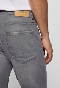 Närmre bild av en person som bär grå denimjeans med bakficka och ett beige läderlappar märkt "BOSS" vid midjan.