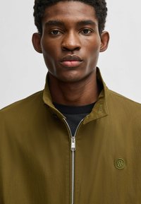Olive green jacket with a zippered front and high collar. Features a small green logo on the chest and textured fabric.