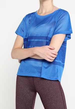 Persoon draagt een blauw mesh sportshirt met korte mouwen over een lichtroze sportbeha en bordeauxrode legging, met de armen gekruist ter hoogte van de taille.