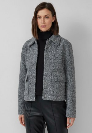 Graue, strukturierte Jacke mit einem Kragenhals, Frontdruckknöpfen und zwei aufgesetzten Taschen. Getragen über einem schwarzen Rollkragenpullover.