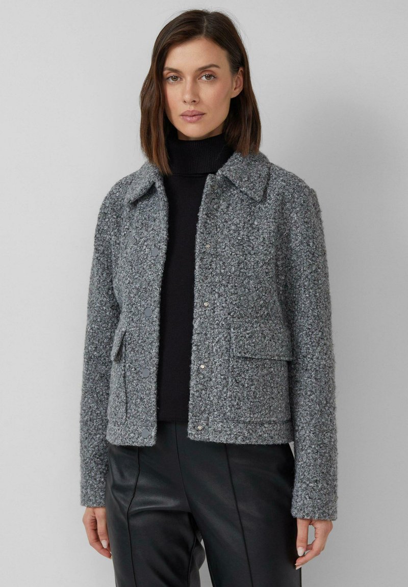 Graue, strukturierte Jacke mit einem Kragenhals, Frontdruckknöpfen und zwei aufgesetzten Taschen. Getragen über einem schwarzen Rollkragenpullover.