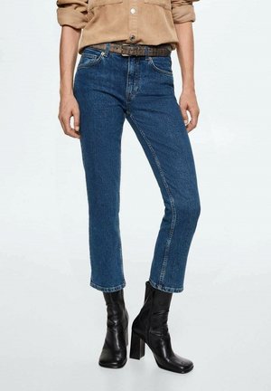 Osoba ubrana w beżową koszulę sztruksową z podwiniętymi rękawami, niebieskie dżinsy o prostych nogawkach z brązowym paskiem oraz czarne botki na obcasie, stojąca na białej podłodze.