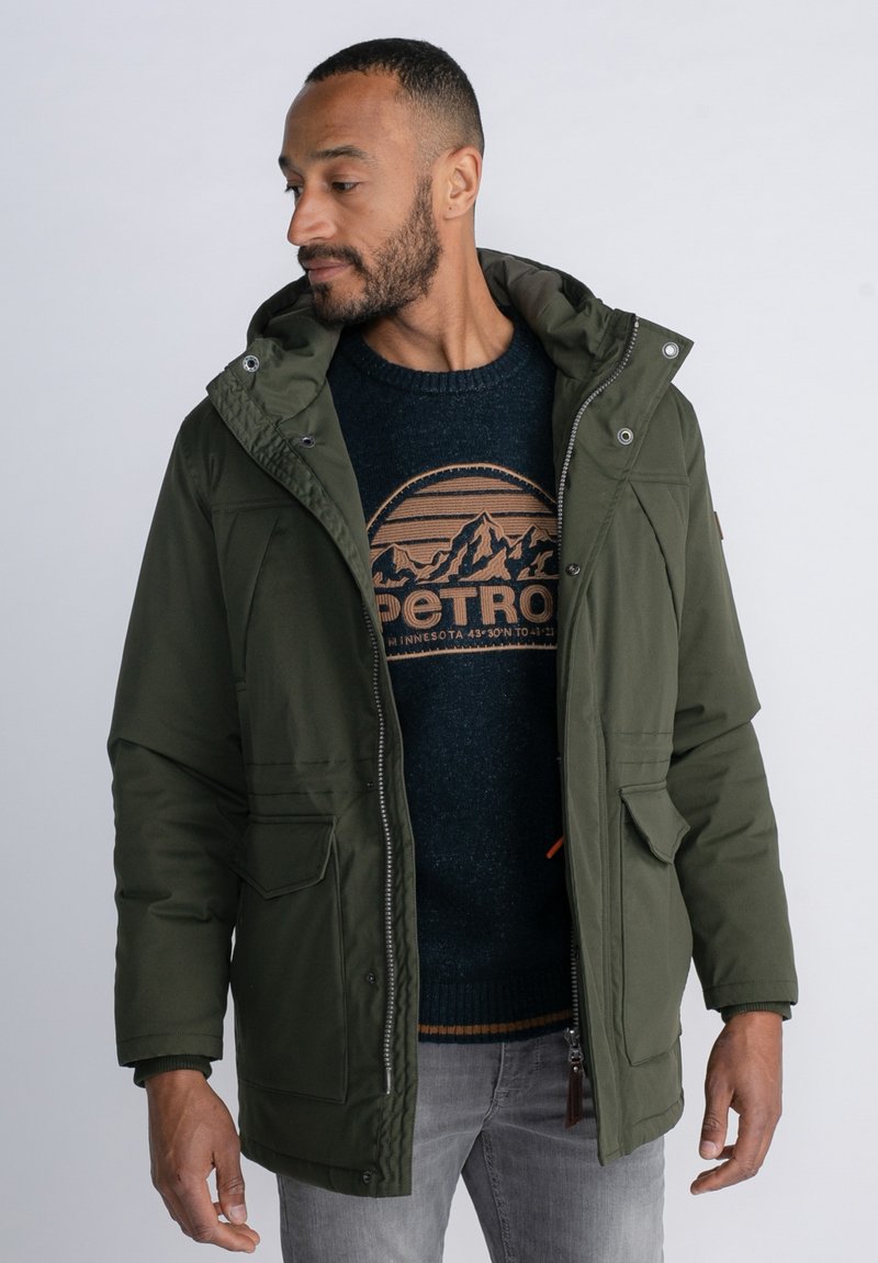 Parka verde oliva con capucha, cremallera y botones a presión. Tiene bolsillos en el pecho y laterales, y se lleva sobre un suéter azul oscuro con un diseño de montañas.