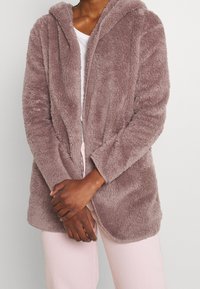 Luddig, mauve huva jacka med öppen framsida, långa ärmar och mjuk textur. Bärs över en vit topp, med armarna korsade för en avslappnad look.