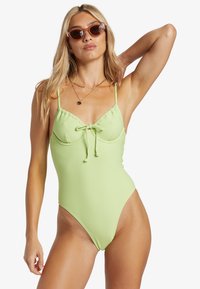Ljusgrön randig baddräkt med justerbara axelband, knytfront och högskurna ben. Modell som bär genomskinliga ovala solglasögon.