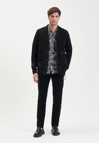 Zwarte gebreide cardigan met knoopsluiting, gedragen over een shirt met patroon. Gecombineerd met zwarte corduroy broek en zwarte instapschoenen.