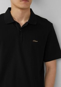 Schwarzes Poloshirt aus Baumwolle mit strukturierten Oberflächen, ausgestattet mit einem Button-Down-Kragen und einem kleinen goldenen, gestickten Logo auf der Brust.