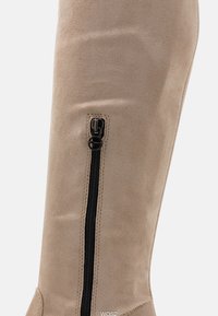 Beige Wildleder-Knöchelstiefel mit einem schwarzen Reißverschlussdetail an der Seite, vertikaler Naht und weicher Textur. Einfaches Design mit minimalen Akzenten.
