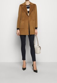 Persona con abrigo color camello, suéter de rayas blanco y negro, pantalones ajustados negros, tacones negros, sosteniendo un bolso acolchado blanco con cadena dorada.