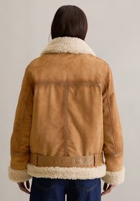 Tan suède jack met een witte schapenvacht kraag en manchetten, voorzien van een ceintuur in de taille en zichtbare stiksels op een gladde textuur.