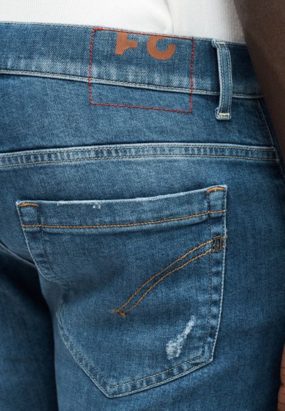 Jean en denim bleu coupe slim, avec une étiquette cousue rouge, deux poches arrière et un détail subtilement usé près de la poche droite.