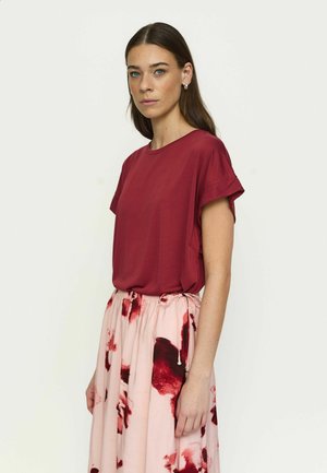 Kvinde med langt mørkt hår iført en kortærmet burgunder top og en lyserød nederdel med røde blomsterprint, stående foran en ensfarvet lys baggrund.