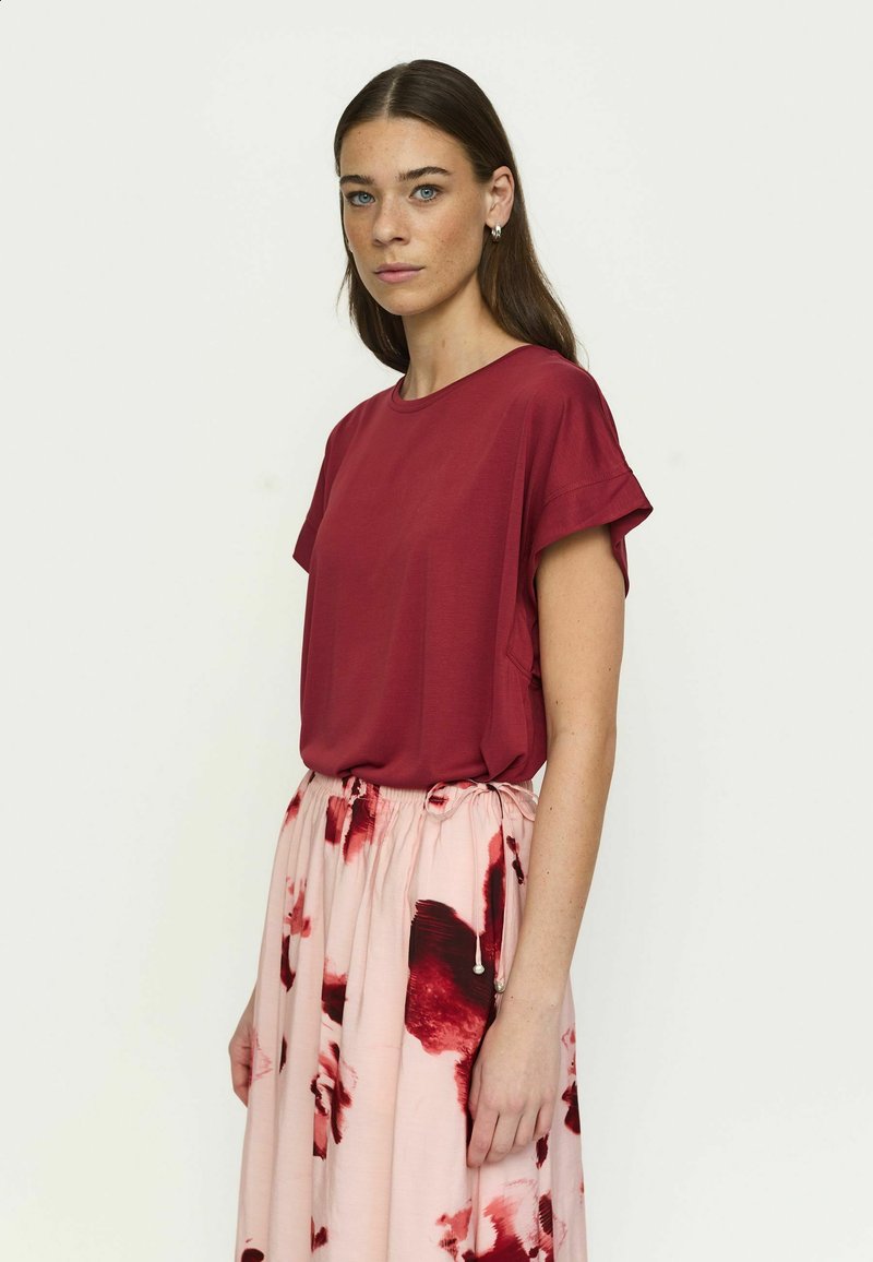 Kvinne med langt mørkt hår har på seg en kortermet burgunder topp og et rosa skjørt med røde blomster, stående foran en ensfarget lys bakgrunn.